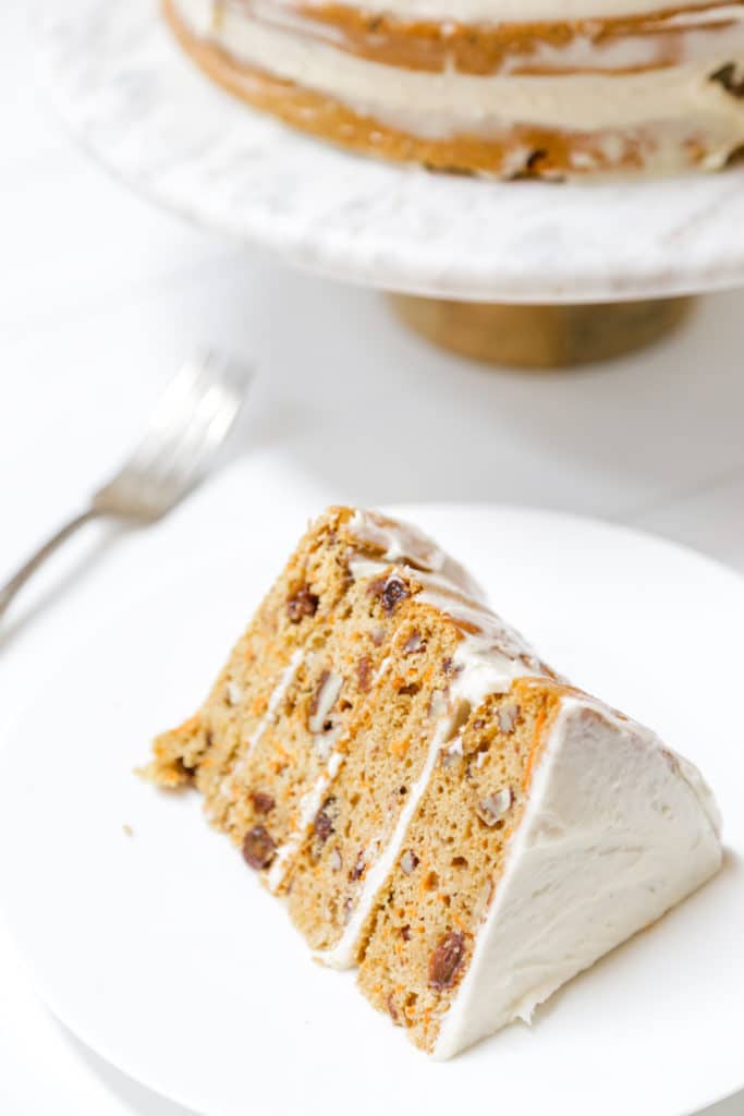 A slice of carrot cake with cream cheese frosting on a white plate with the whole cake in the background.