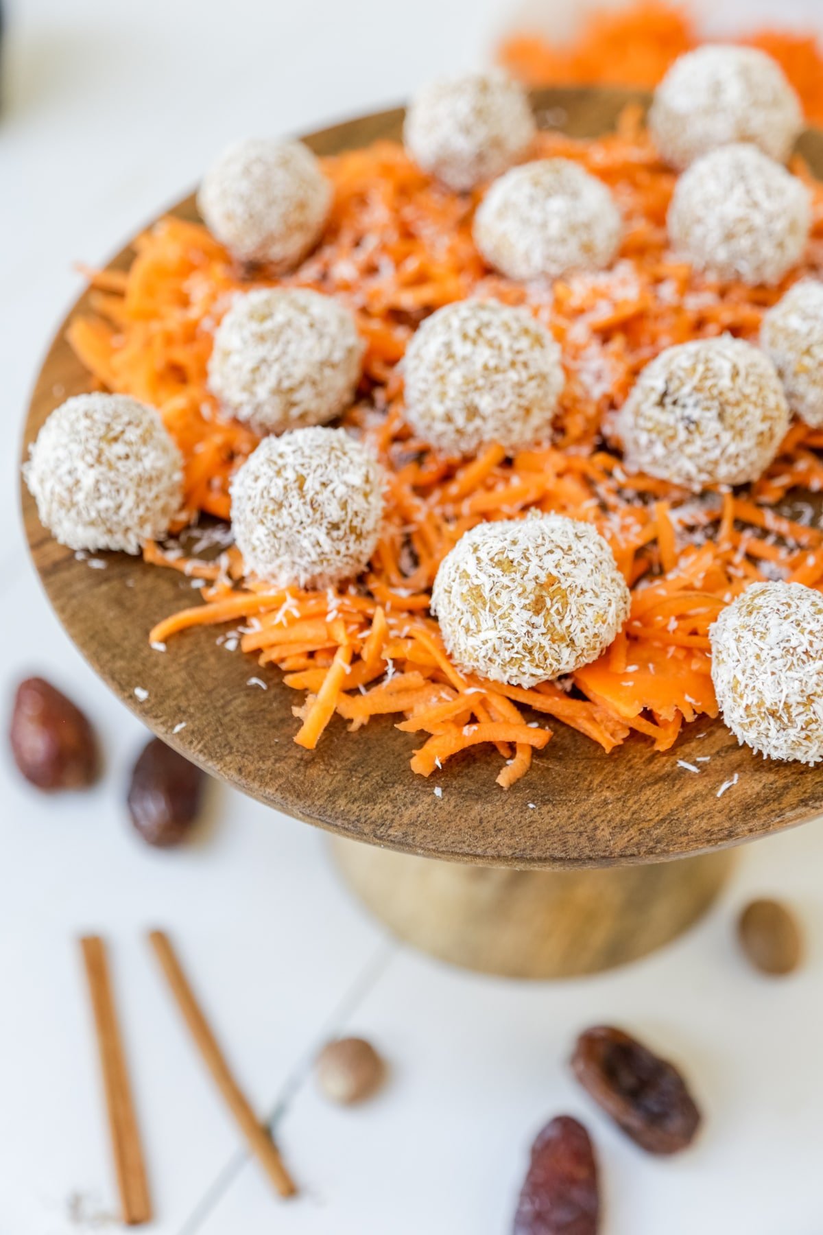 Carrot cake balls rolled in coconut sitting on grated carrots on a wooden cake stand.