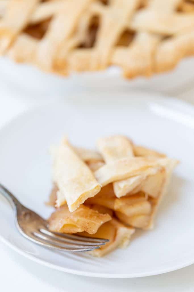 A slice of apple pie with a silver spoon on a white plate and a lattice top apple pie in the background.