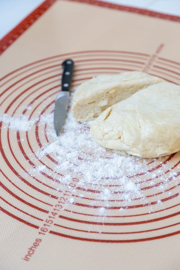 A silicone pie dough mat with a ball of dough cut in half sitting on flour.