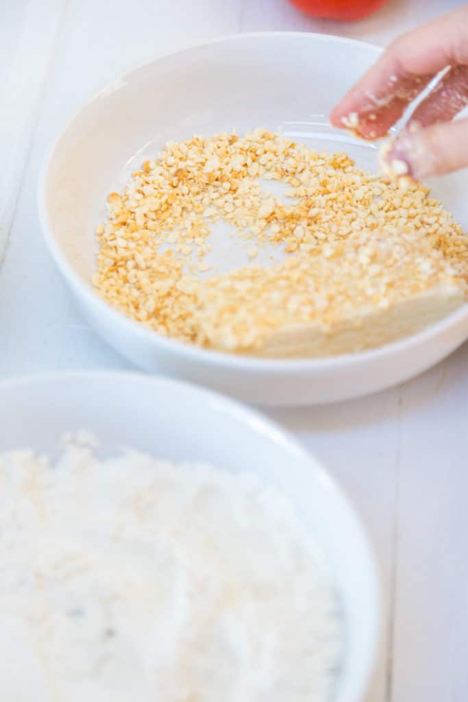 A hand dredging a piece of tofu through breadcrumbs.