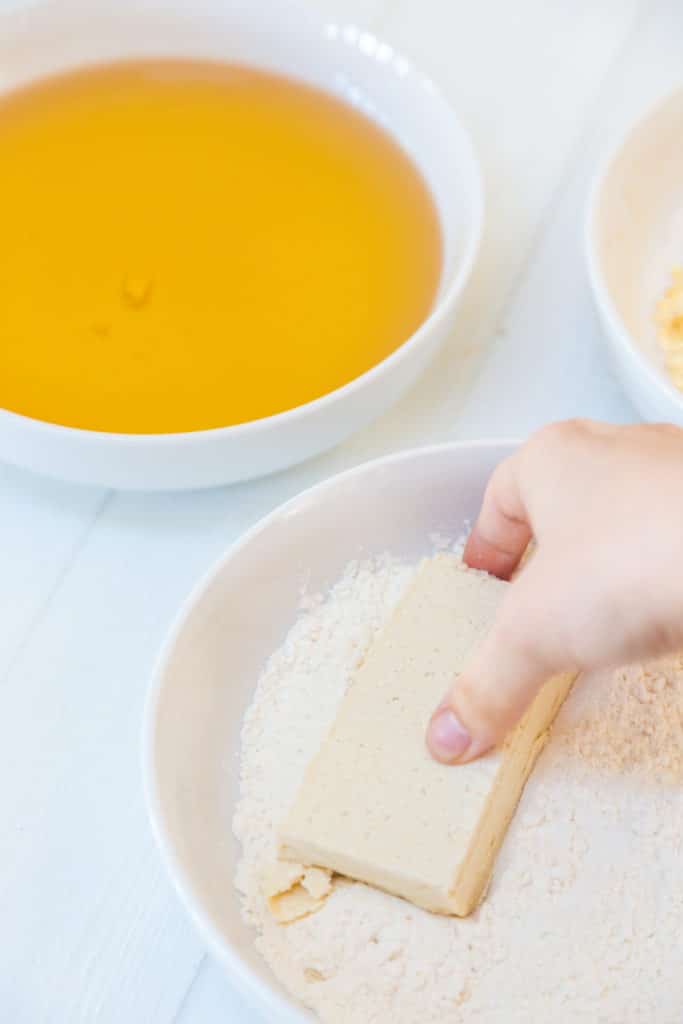 A hand dredging a slice of tofu through flour.