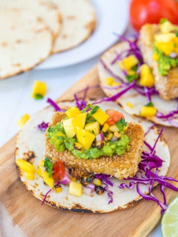 Breaded tofu tacos with mango salsa on a wooden board with flour tortillas in the background.