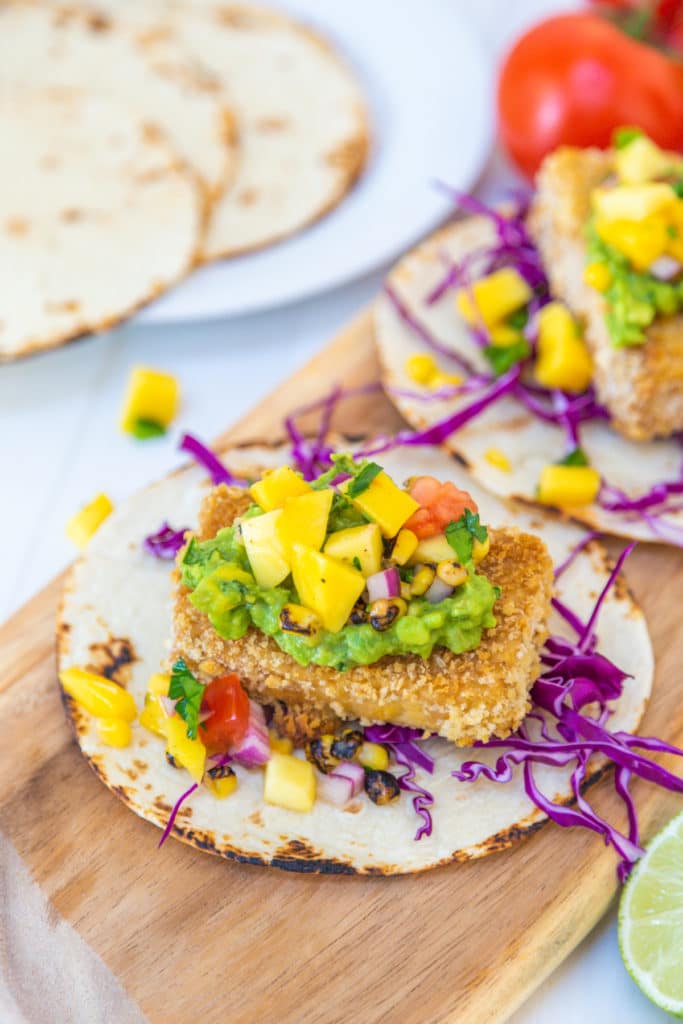 Breaded tofu tacos with mango salsa on a wooden board with flour tortillas in the background.