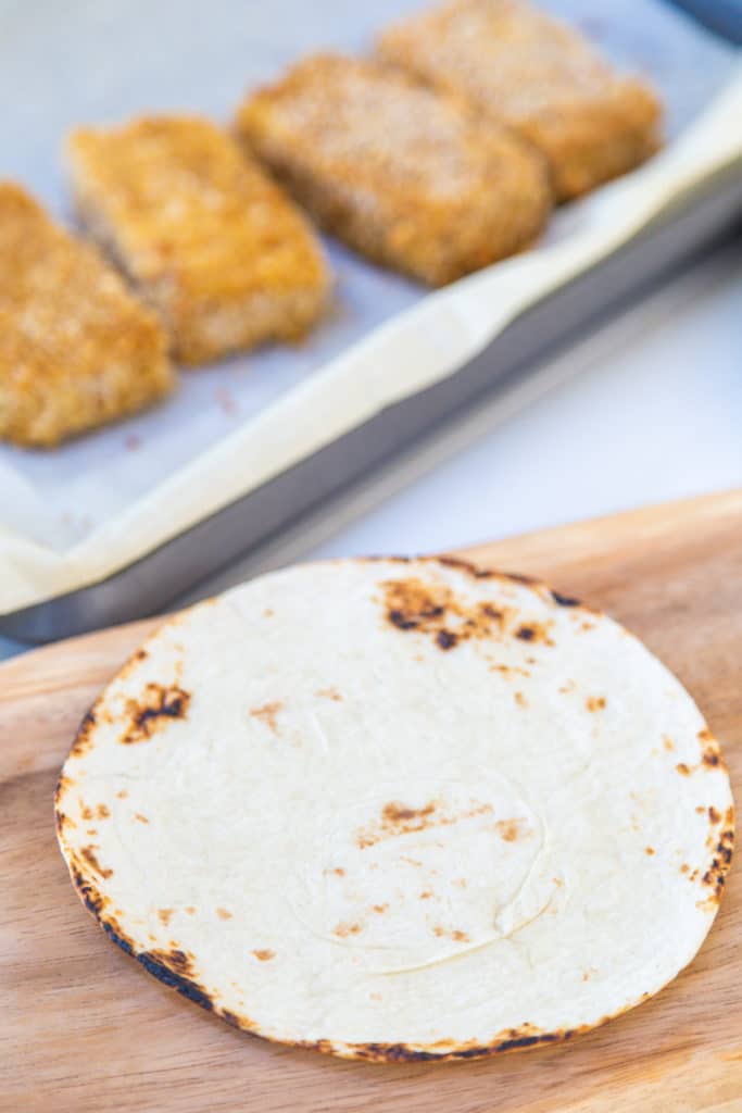 A flour tortilla on a wooden board with fried breaded tofu on a plate.