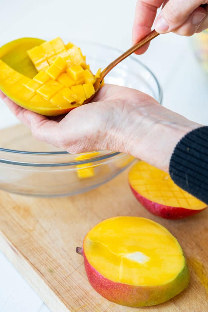 A hand holding a gold spoon that's scooping out cubed mango into a bowl. 