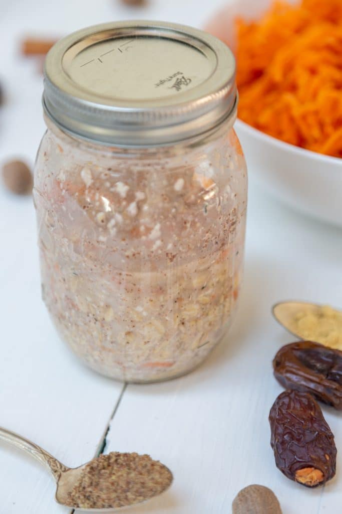 Carrot cake overnight oats in a mason jar with dates, spices, and a bowl of shredded carrots next to the jar.