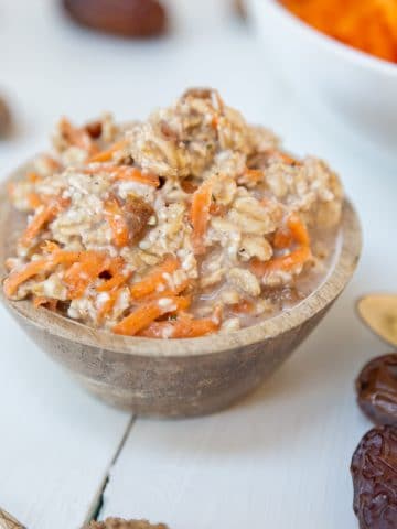 Carrot cake overnight oats in a wooden bowl with dates, spices, and shredded carrots surrounding the bowl.