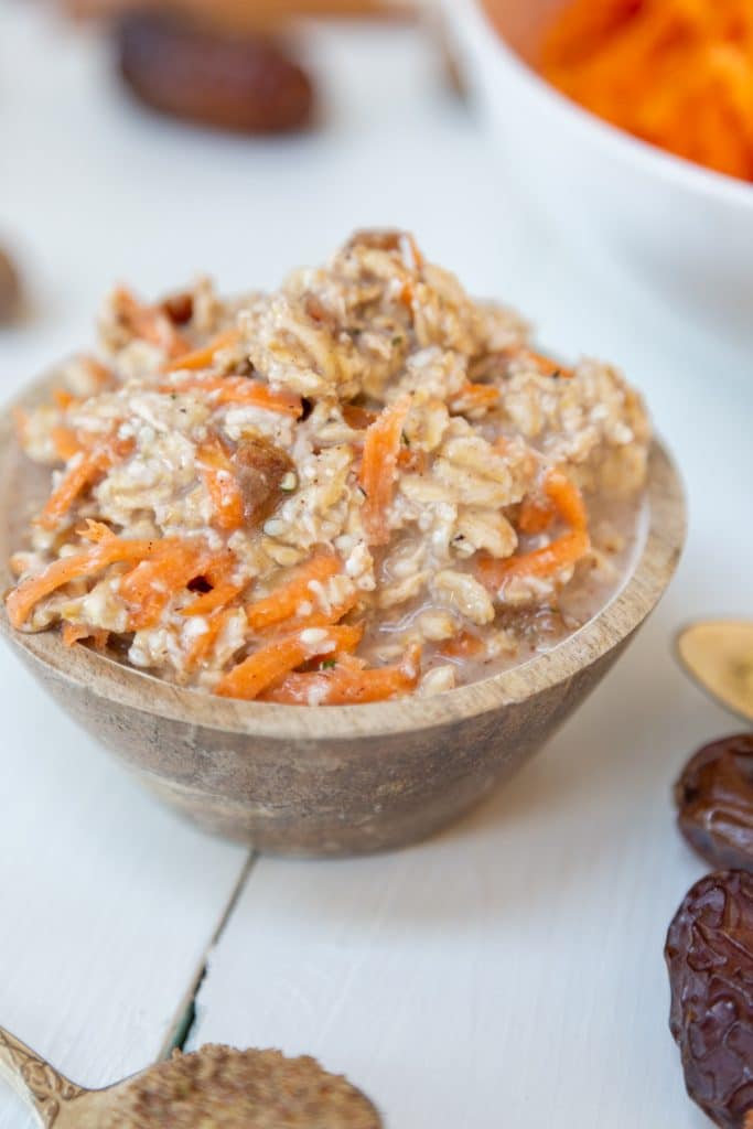 Carrot cake overnight oats in a wooden bowl with dates, spices, and shredded carrots surrounding the bowl.