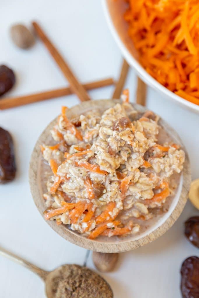 An overhead shot of a wooden bowl filled with carrot cake overnight oats and a corner of a white bowl with shredded carrots, and cinnamon sticks and dates around the bowl.