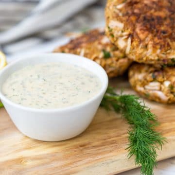 A white bowl of tartar sauce and crab cakes on a wooden board with a sprig of fresh dill.