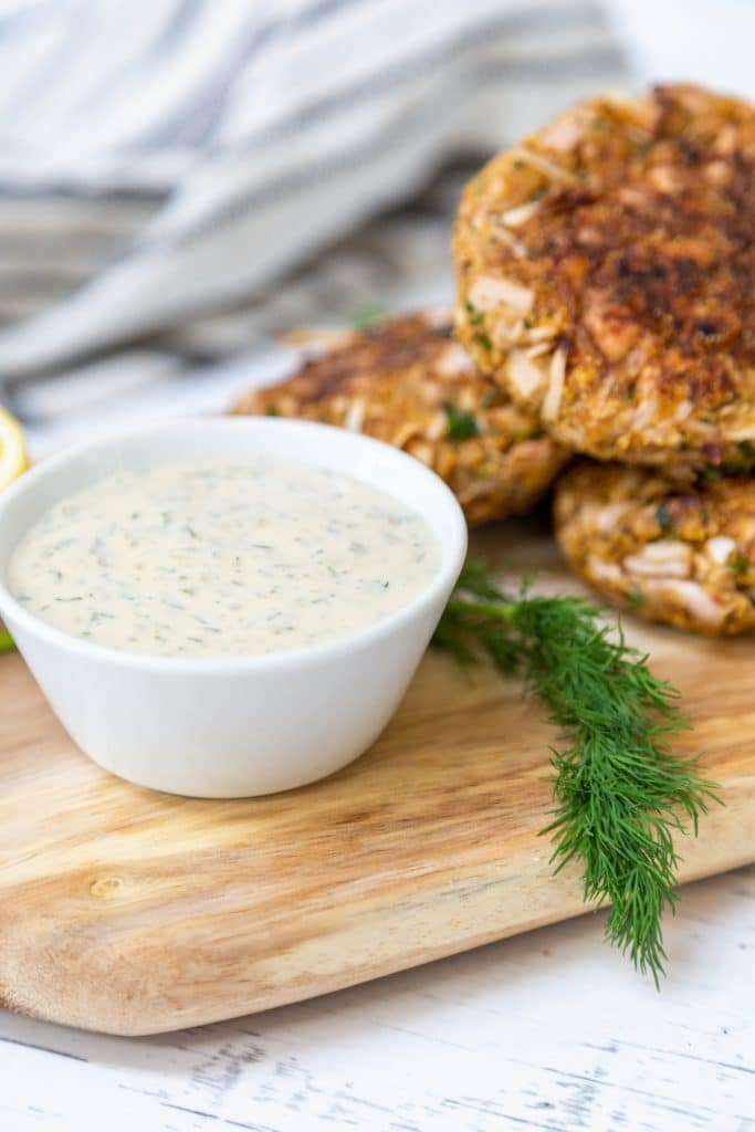 A white bowl of tartar sauce and crab cakes on a wooden board with a sprig of fresh dill.