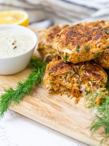 Crab cakes piled on a wooden board with a bowl of tartar sauce and dill.