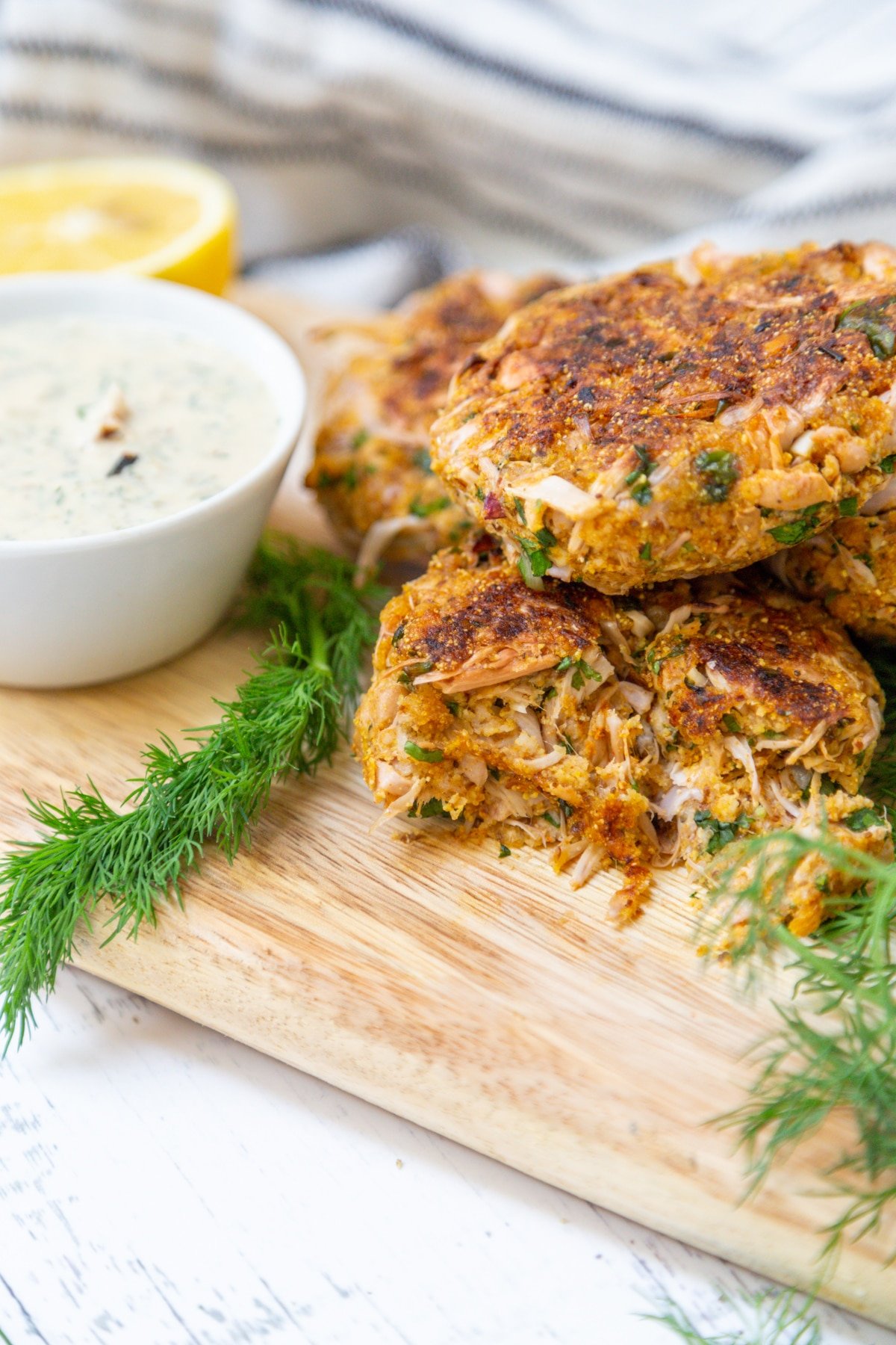 Crab cakes piled on a wooden board with a bowl of tartar sauce and dill.