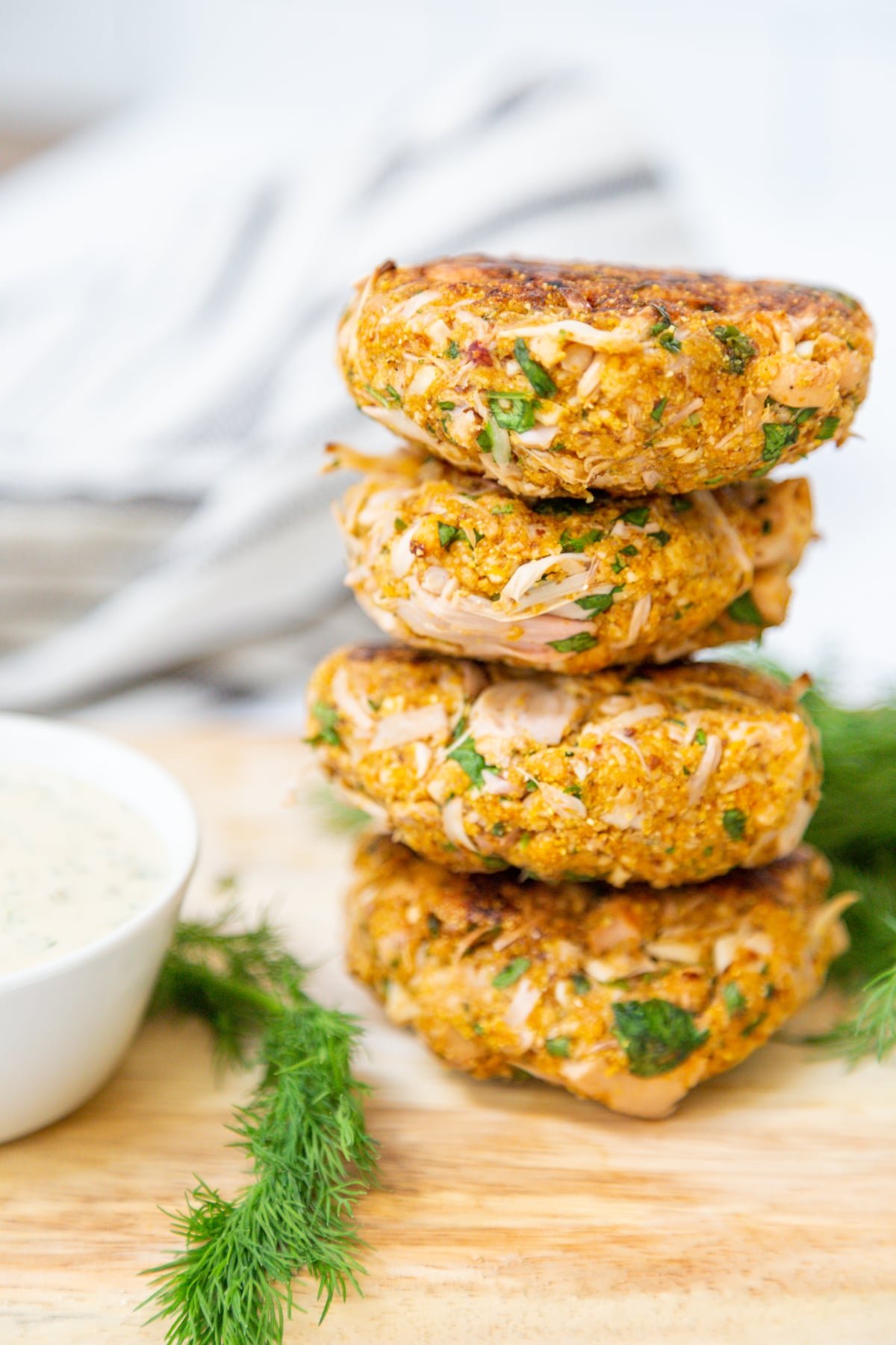 4 crab cakes stacked on a wooden board with dill and a bowl of tartar sauce.