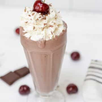 A chocolate milkshake in a tall glass with whipped topping and a cherry on top.