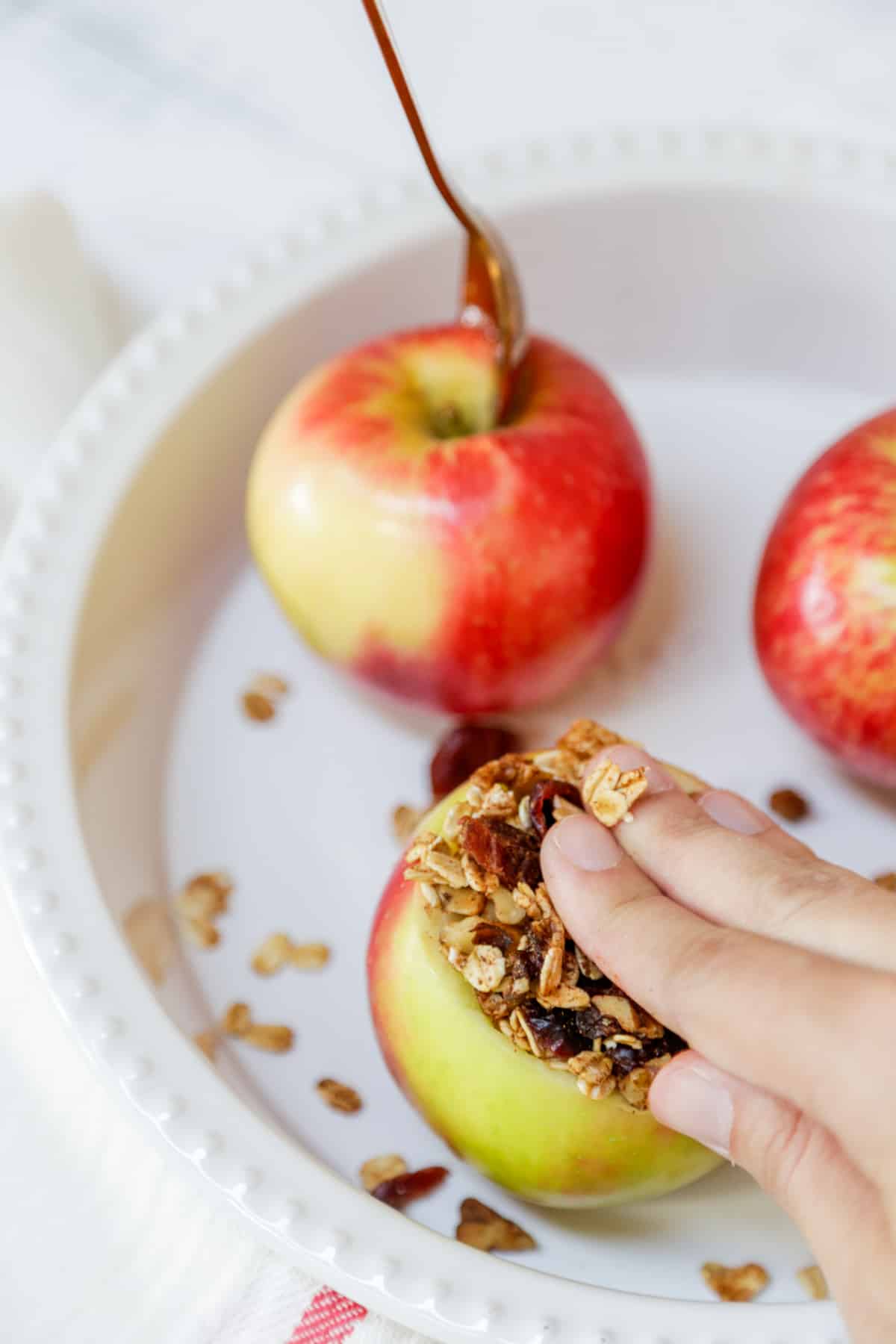 A hand pressing oat filling into a cored apple with whole apples next to the stuffed apple.
