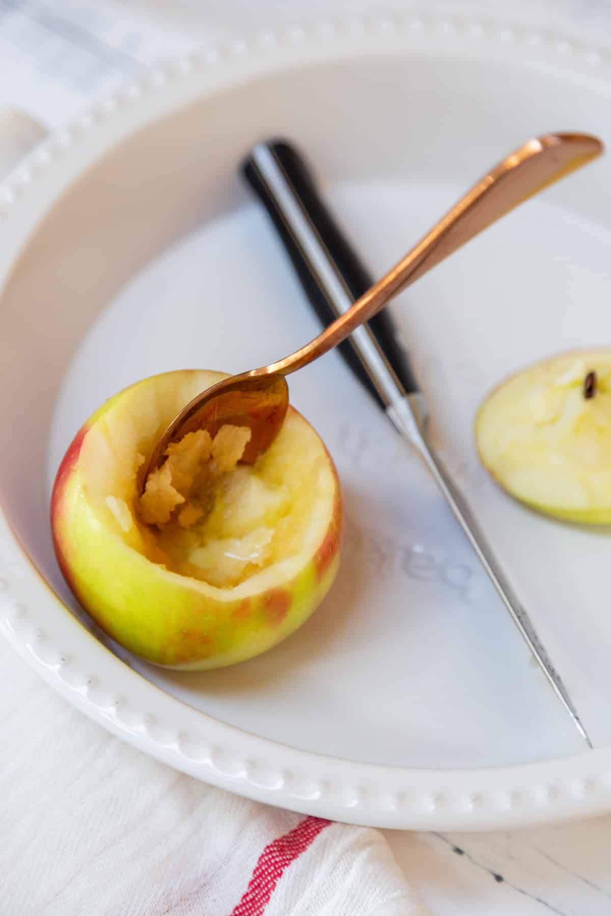 An apple with the core removed in a white baking dish with a gold spoon in the apple and a paring knife next to the apple.