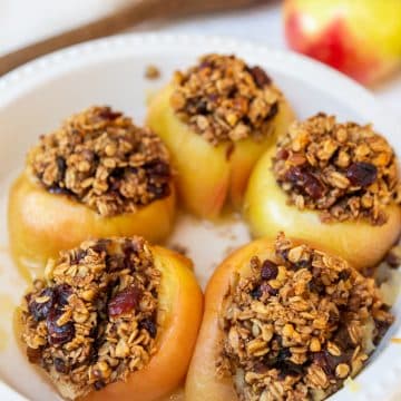 A round white baking dish with 5 stuffed baked apples and a wooden spoon and raw red apples in the background.