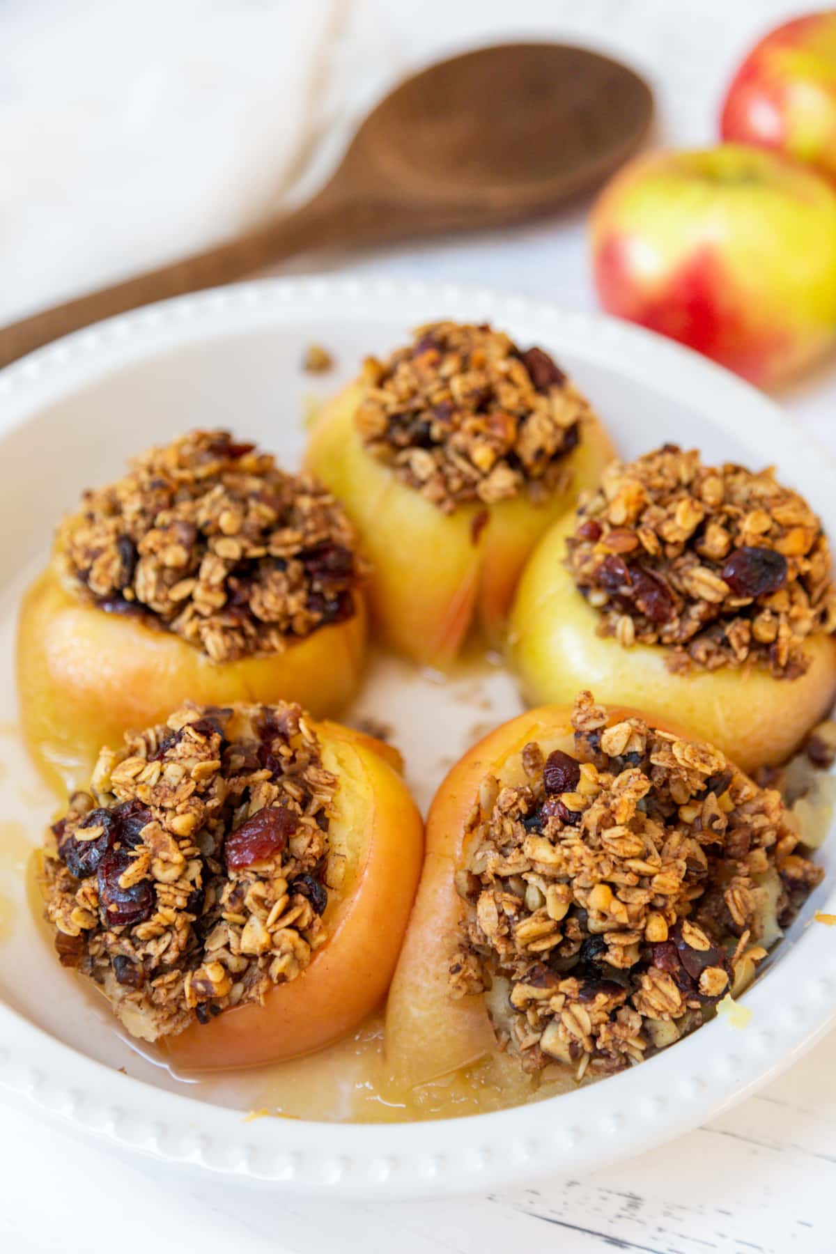 A round white baking dish with 5 stuffed baked apples and a wooden spoon and raw red apples in the background.