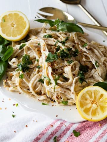 A white platter with fettuccini Alfredo sprinkled with parsley and lemon halves on the side of the plate.