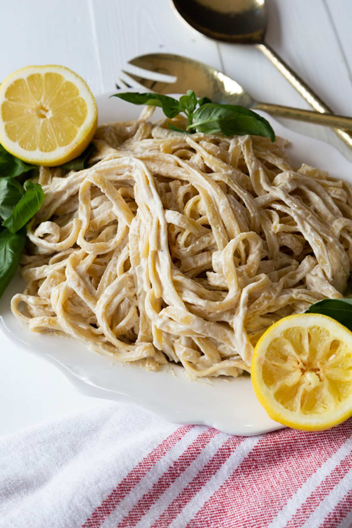 A white platter of fettuccini Alfredo with fresh basil and lemon halves.