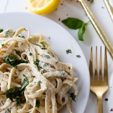 A white plate with vegan fettuccine Alfredo sprinkled with chopped basil and a gold fork and serving utensils next to the plate.