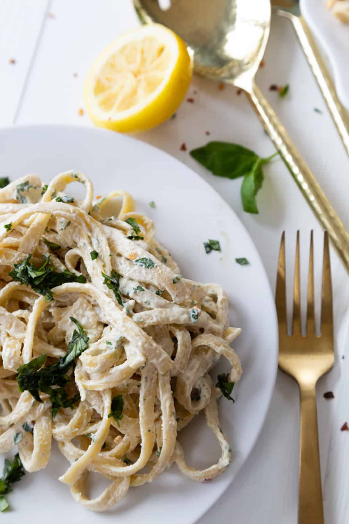 A white plate with vegan fettuccine Alfredo sprinkled with chopped basil and a gold fork and serving utensils next to the plate.