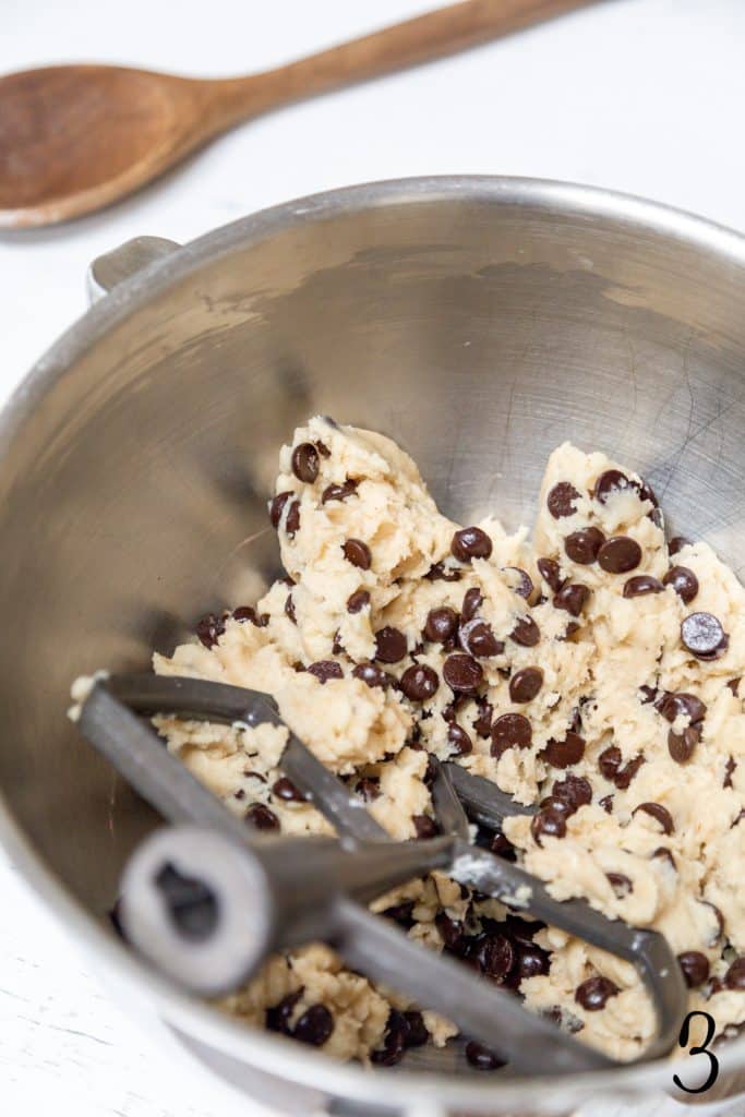 Chocolate chip cookie dough in a silver mixing bowl with a paddle attachment.