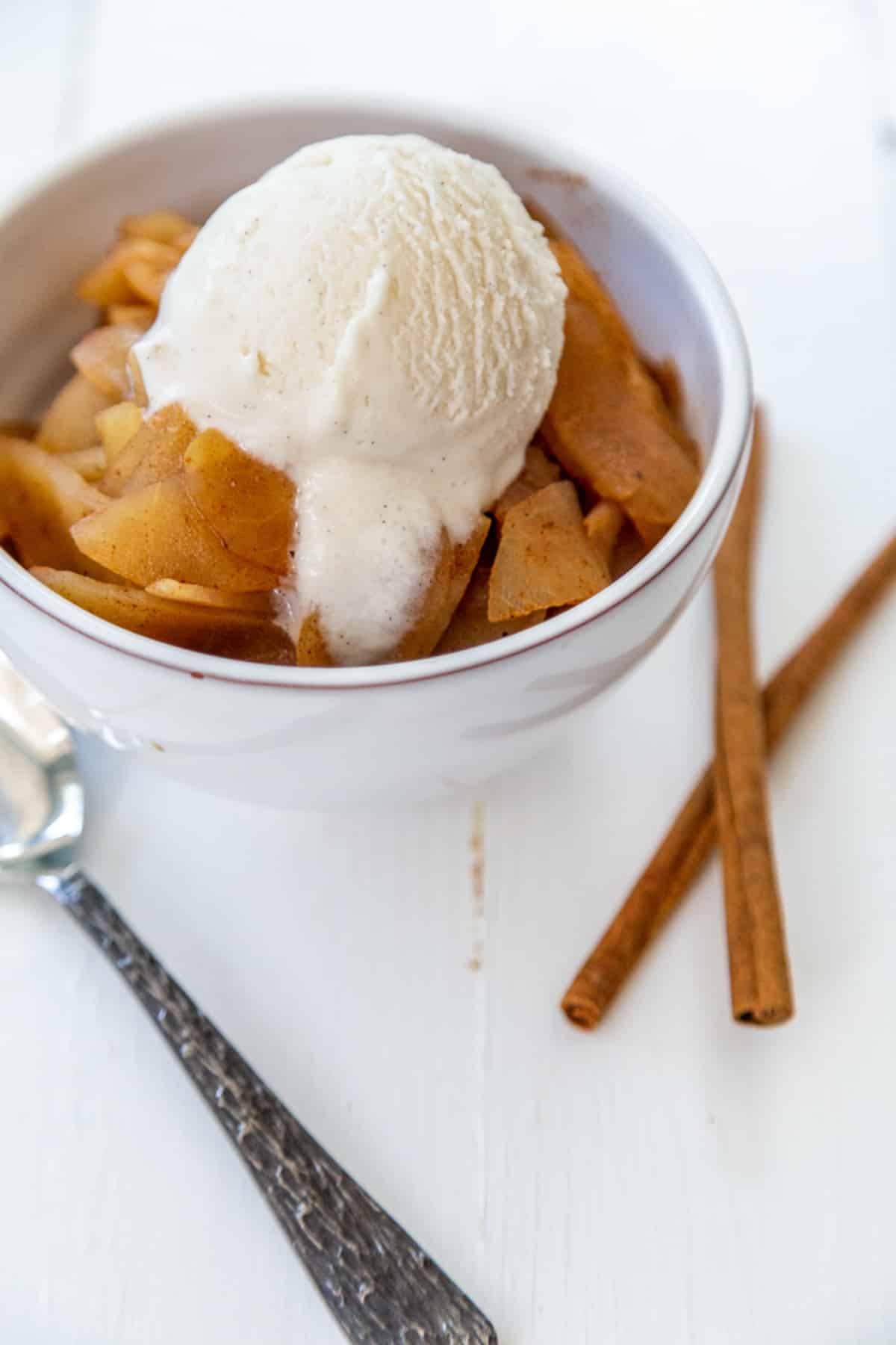 A white bowl of chunky applesauce with a scoop of vanilla ice cream on top and 2 cinnamon sticks and a spoon next to the bowl.