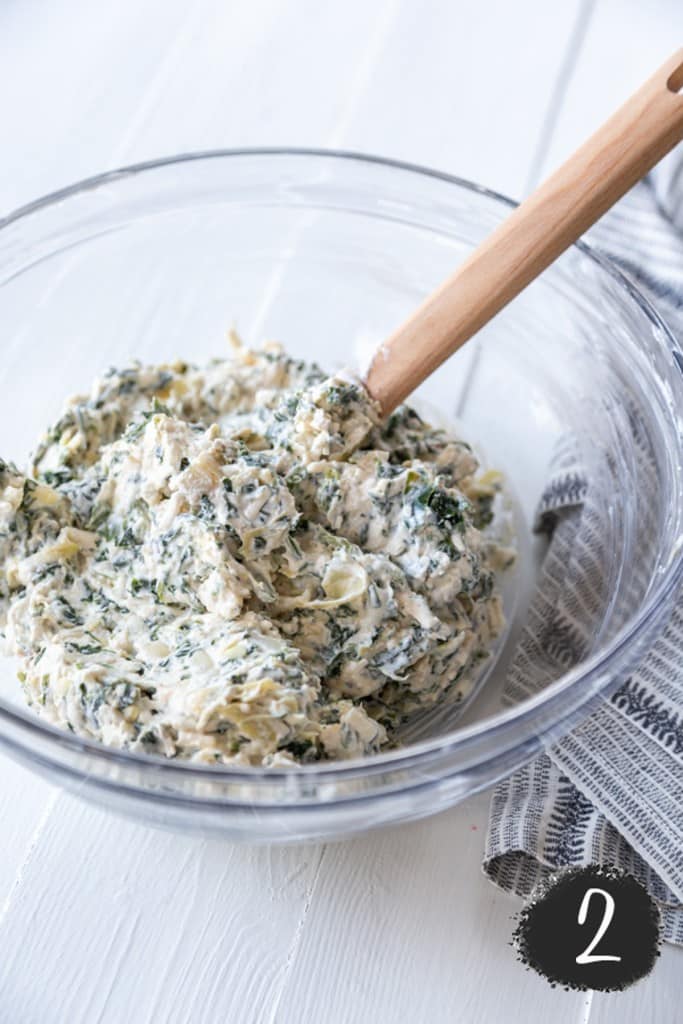 A clear mixing bowl with spinach artichoke dip being stirred with a wooden spoon. 
