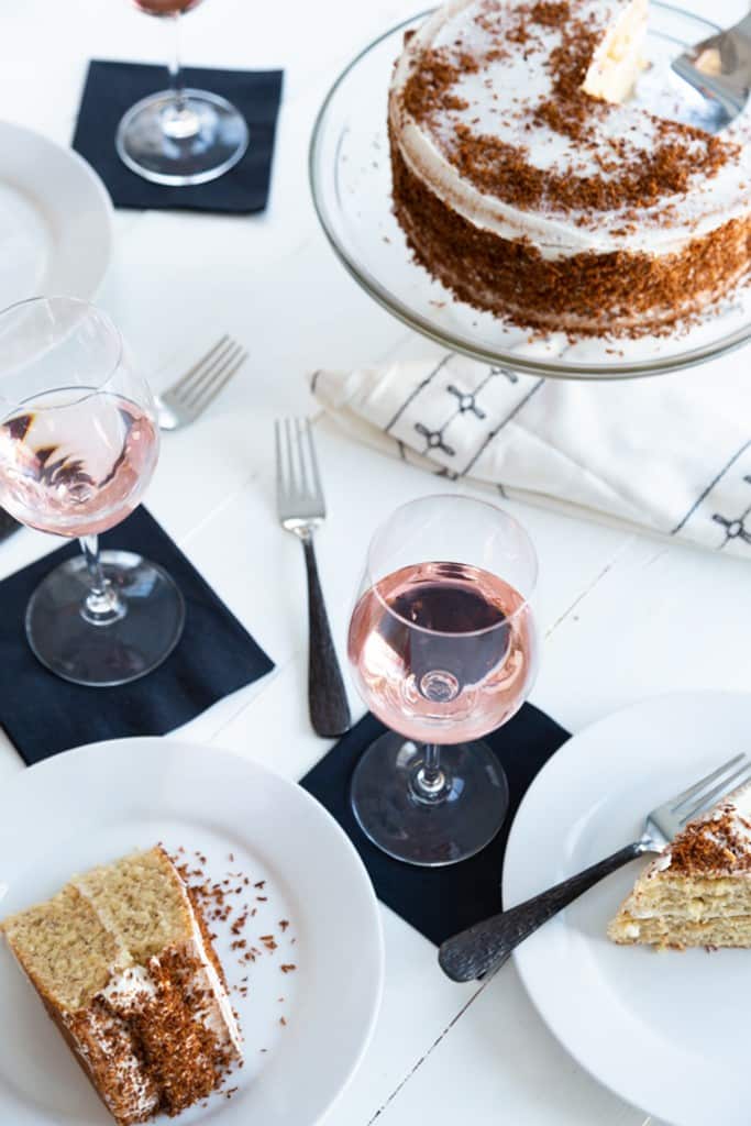 A white table with a layer cake with vanilla frosting and white plates with slices of the cake and rose wine in glasses.
