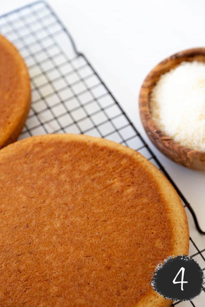 Two round yellow layer cakes on a wire cooling rack with a wooden bowl of coconut next to the cake.