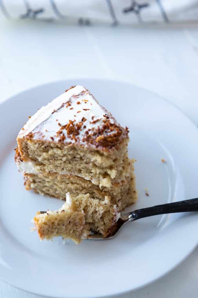 A slice of banana cake on a white plate with a forkful of the cake on the plate.