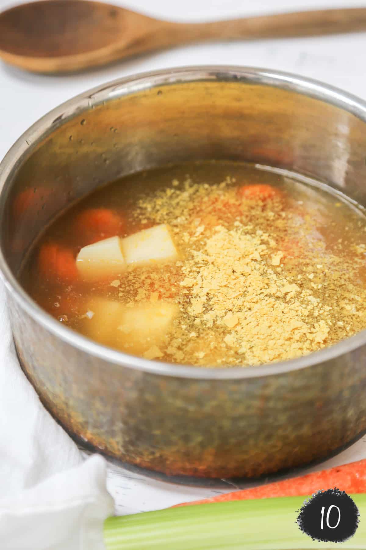 A copper pot with nutritional yeast floating on top of broth, potatoes, and carrots.