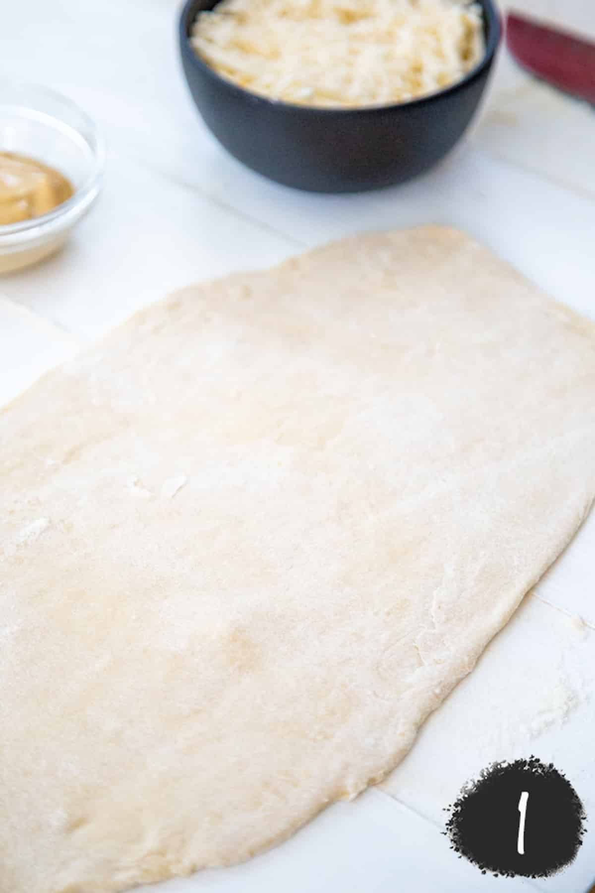 Pizza dough rolled out on a marble board with a black bowl of shredded white cheese and a rolling pin next to the dough.