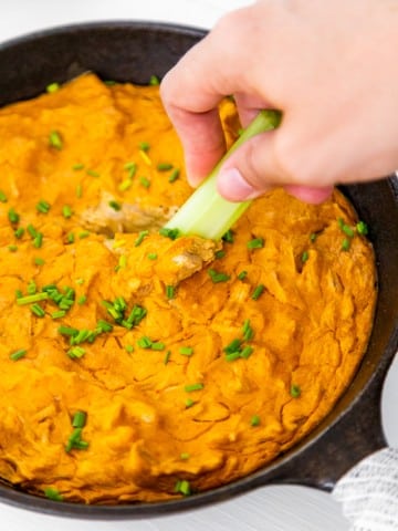 An iron skillet with buffalo chik'n dip and a hand dipping a stalk of celery in it.