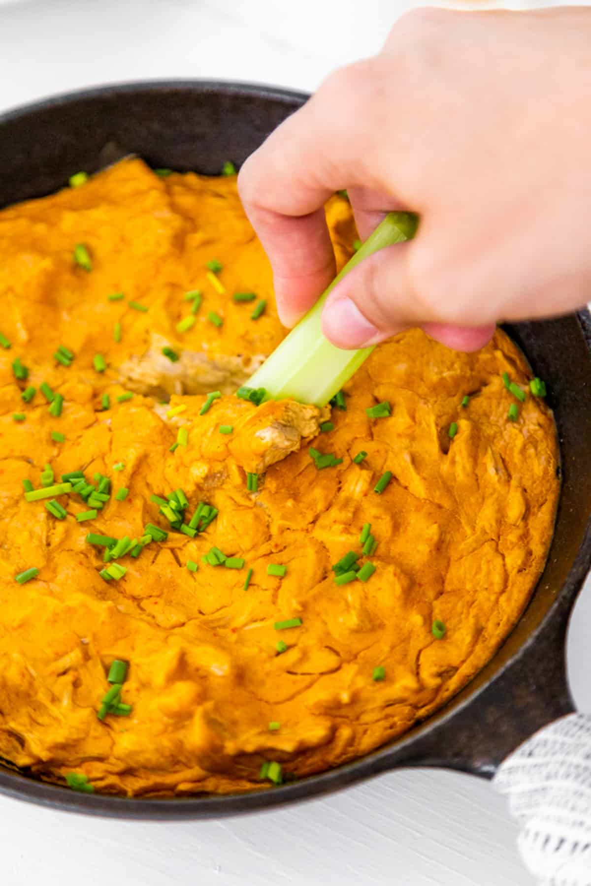 An iron skillet with buffalo chik'n dip and a hand dipping a stalk of celery in it.