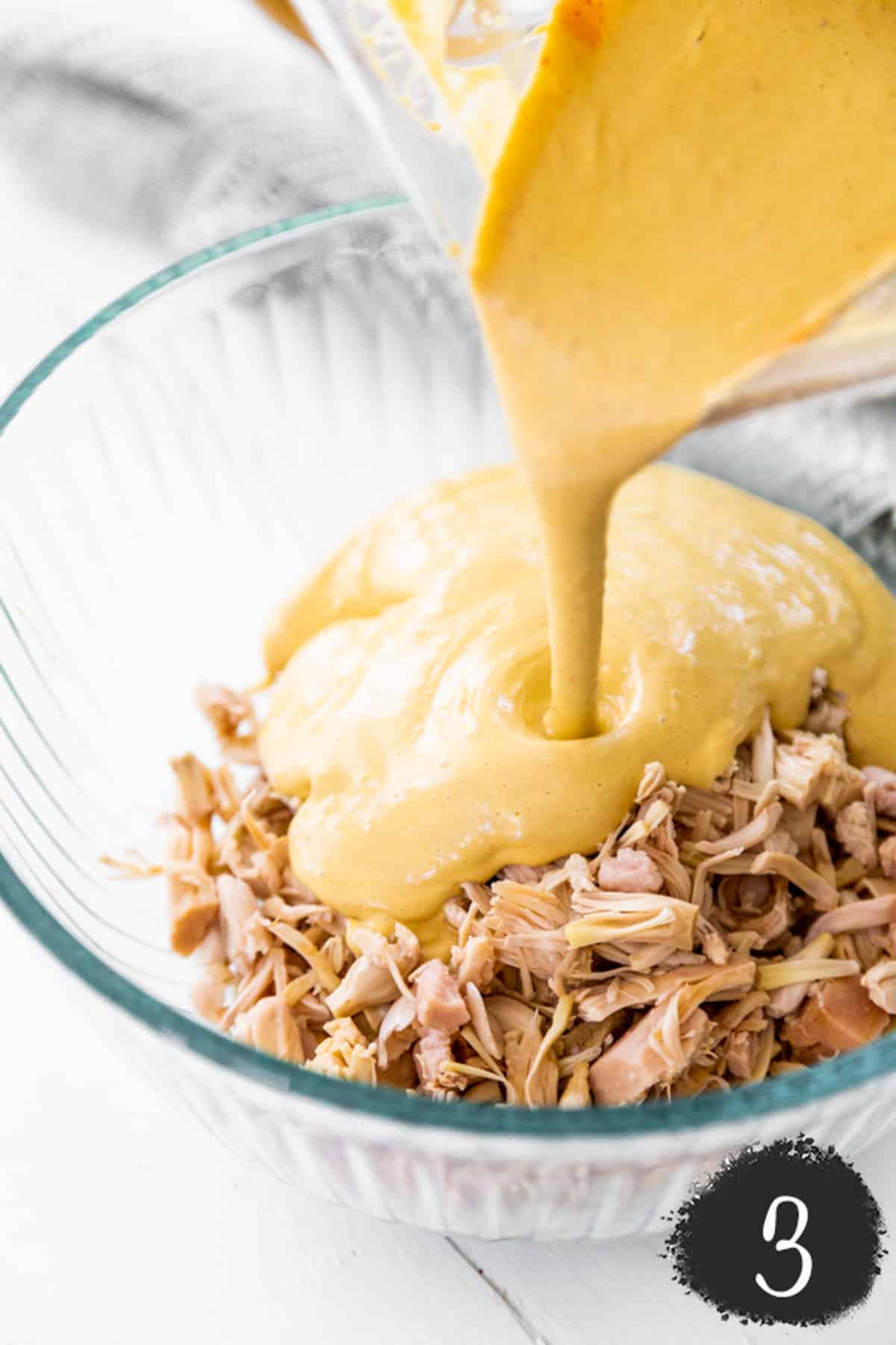 Shredded jackfruit in a large clear mixing bowl and buffalo dip sauce is being poured over the jackfruit.