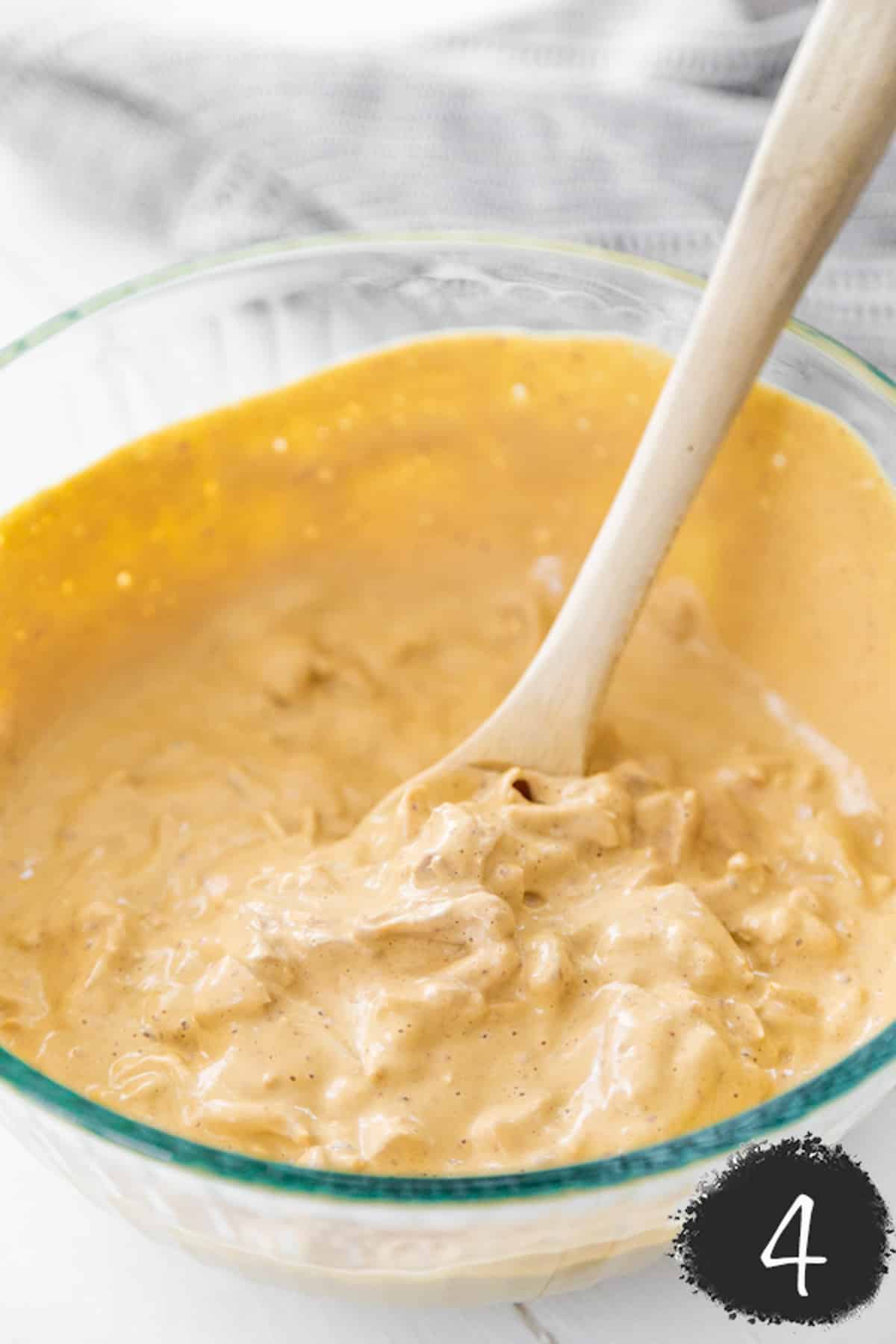 A spoon stirring shredded jackfruit and a creamy buffalo sauce together in a large glass mixing bowl.