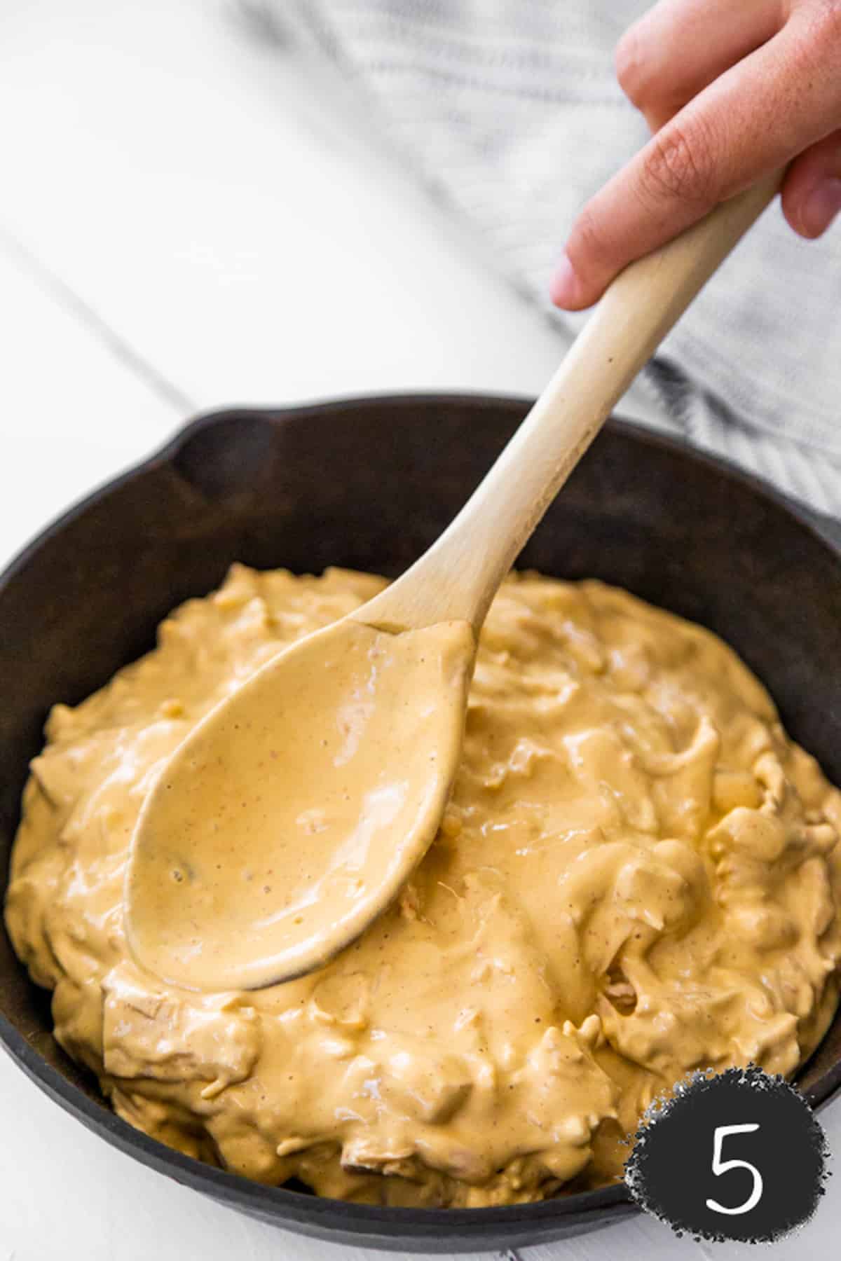A hand holding a wooden spoon spreading buffalo chicken dip evenly into a cast iron skillet.