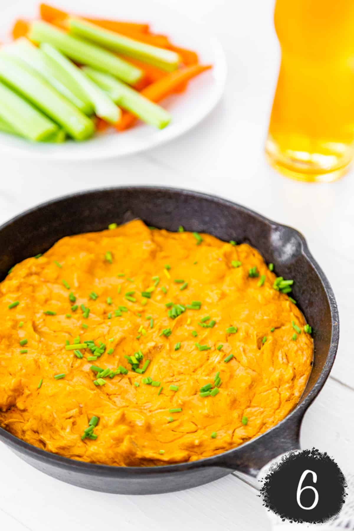 Baked buffalo chicken dip in a cast iron skillet with a white plate of celery and carrot sticks and a glass of beer next to the skillet.