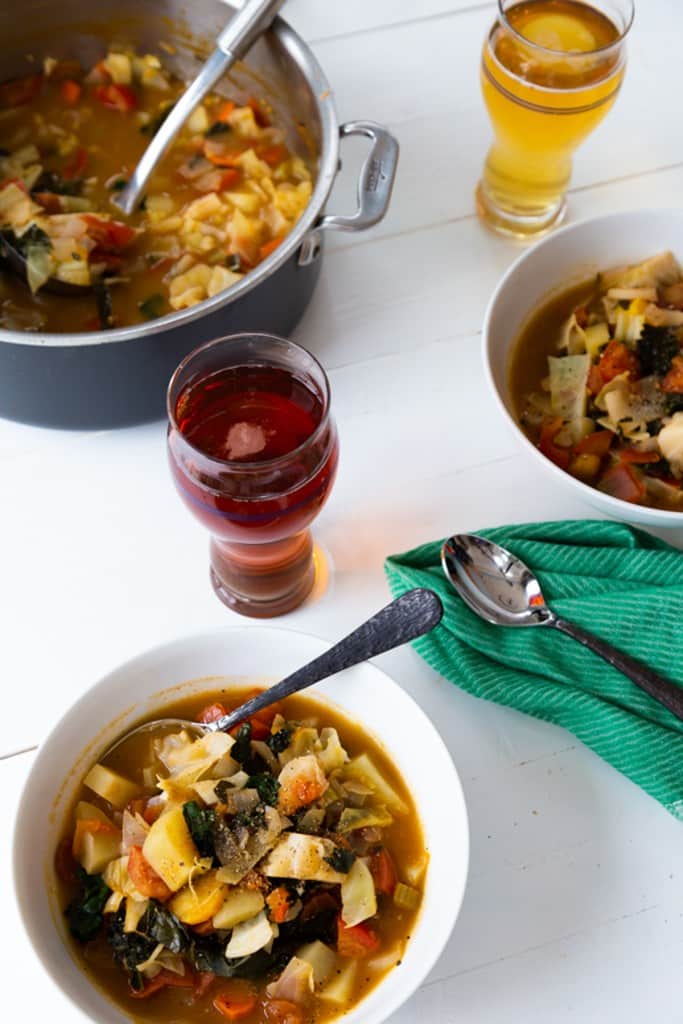 A white table with a white bowl of vegetable soup and a pot of the soup and another bowl of soup and 2 glasses of beer next to it.