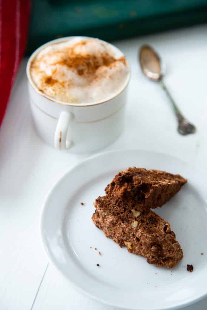 Two pieces of chocolate biscotti on a white plate with a cup of coffee and foamy milk sprinkled with cinnamon next to it and a small silver spoon next to the cup on a white table