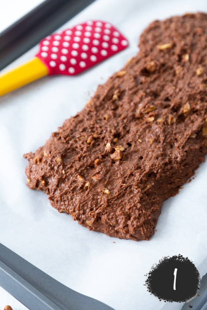 Chocolate biscotti dough molded into a log on a cookie sheet lined with parchment paper and a red and white polka dot spatula with a yellow handle next to it.
