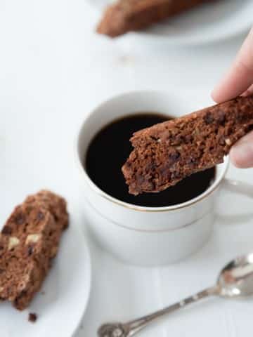 Chocolate chip and pecan biscotti being dipped into a cup of coffee in a white cup with a white plate of biscotti next to it and a silver spoon on the white wood table