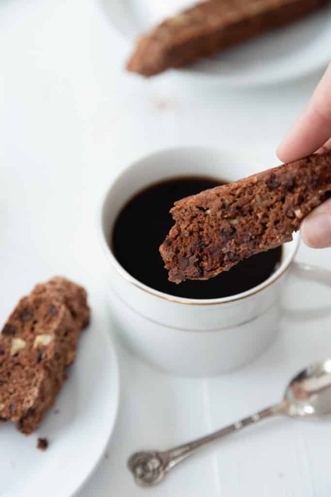 Chocolate chip and pecan biscotti being dipped into a cup of coffee in a white cup with a white plate of biscotti next to it and a silver spoon on the white wood table