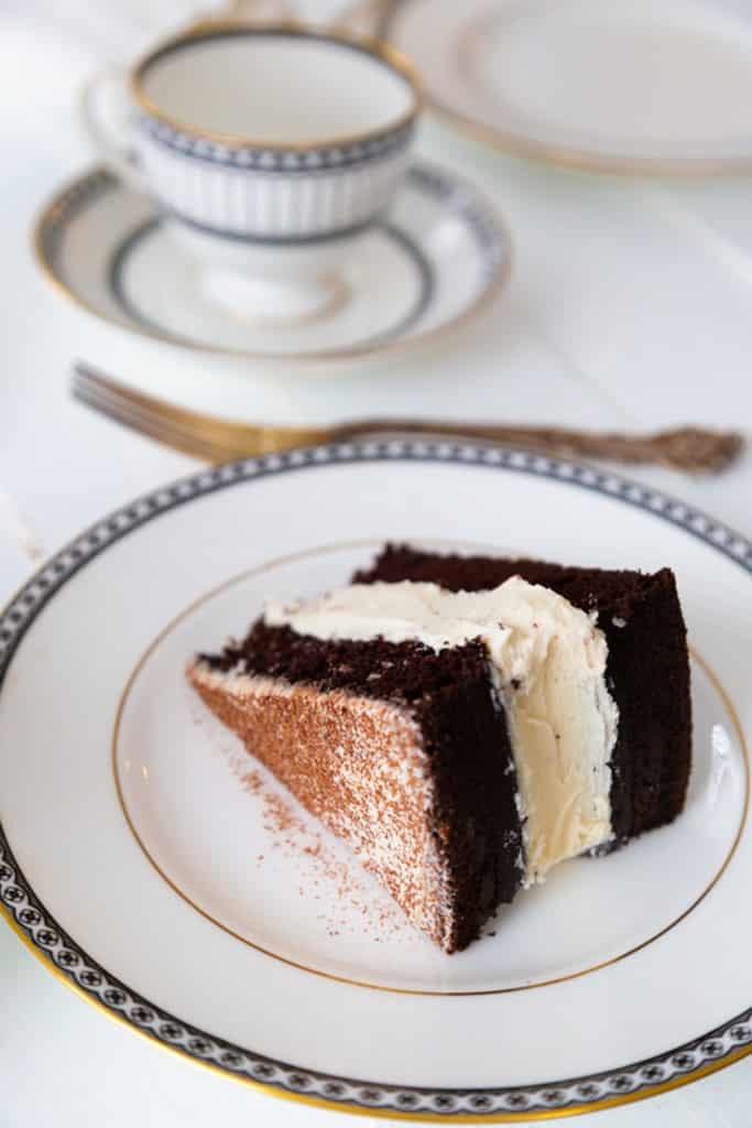 A slice of chocolate layer cake with buttercream frosting on a white plate with a tea cup in the bakground.