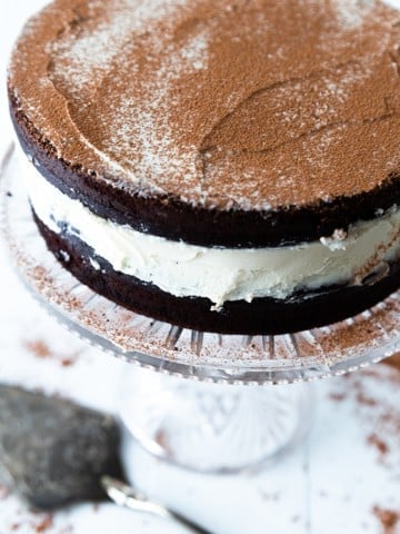 A chocolate cake with white frosting on a glass cake plate with a silver cake knife next to it.