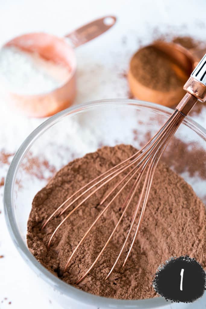 A clear mixing bowl with flour and cocoa powder being whisked with a copper whisk.